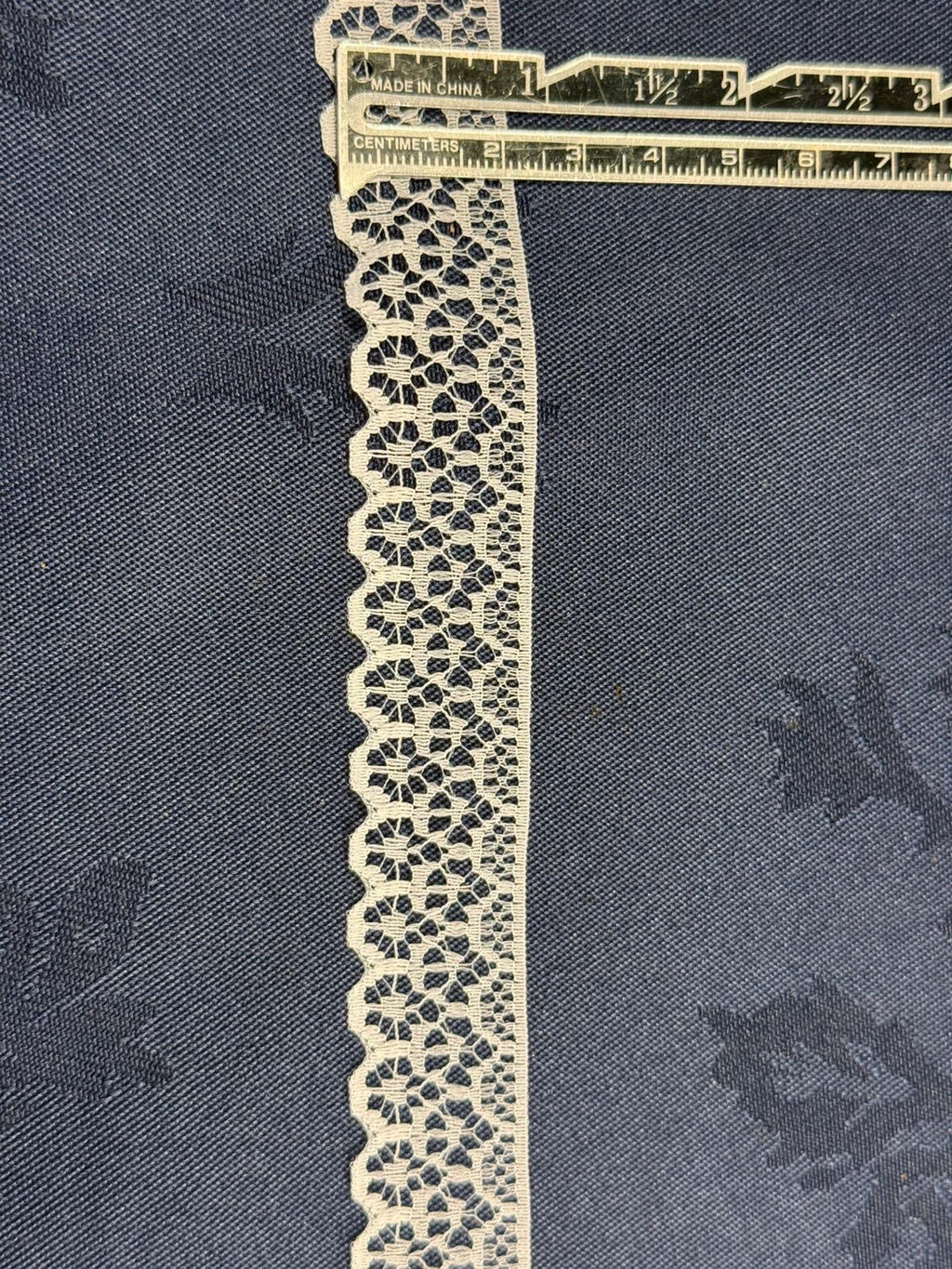 Ivory flat lace trim 25mm wide with scalloped edge and open geometric pattern, shown on denim fabric with a ruler for scale