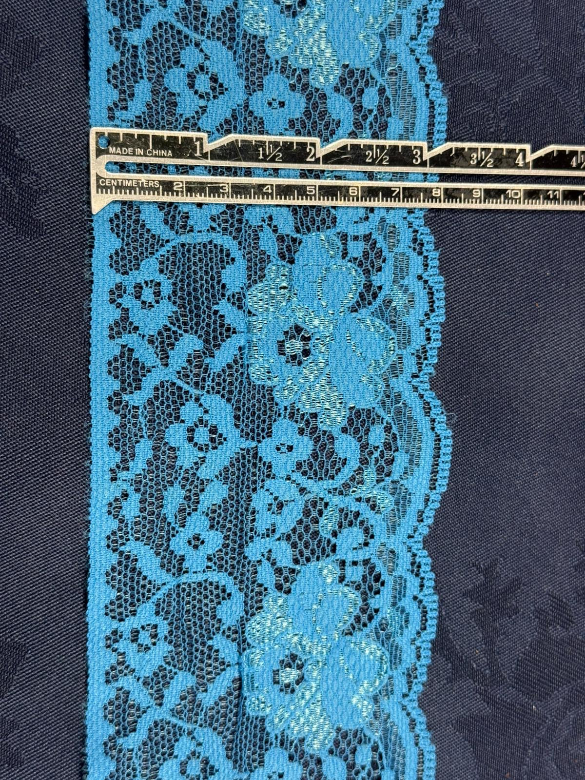 Turquoise flat lace trim 80mm wide with floral scroll pattern and scalloped edge, shown on denim fabric with a ruler for scale